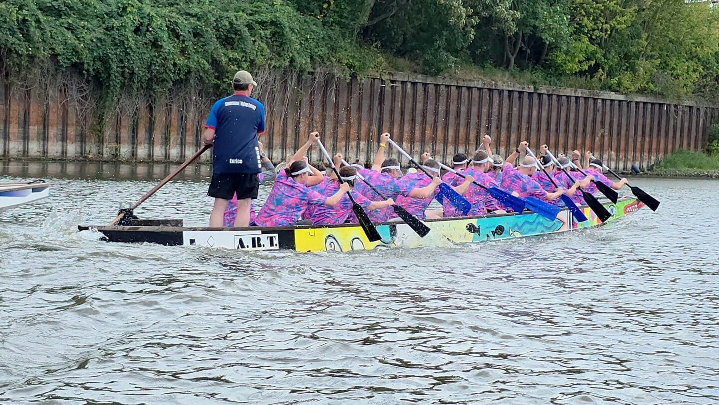 Ein großes buntes Boot mit mehreren Menschen in blauen Trikots die auf einem Fluss paddeln.