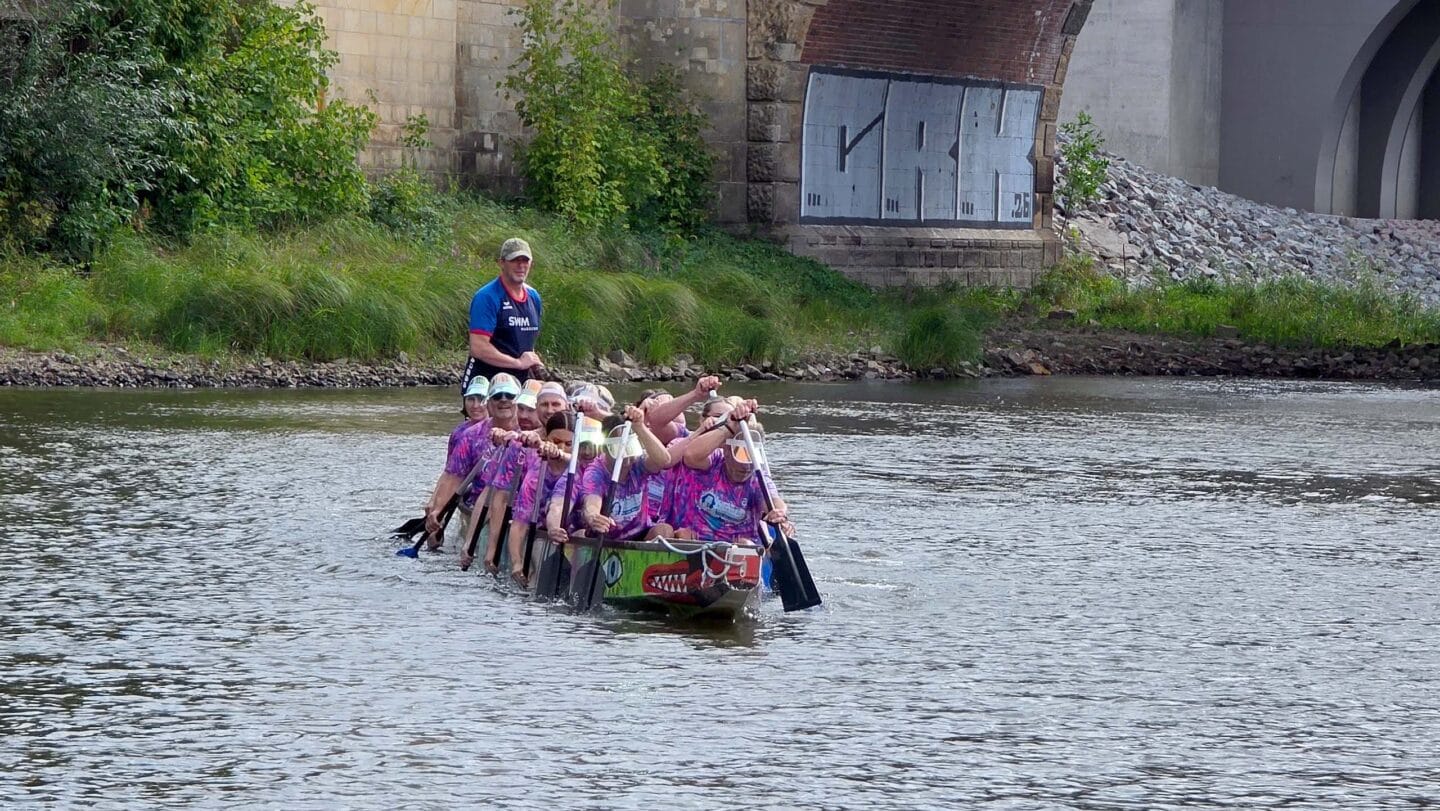 Ein großes weißes Boot mit mehreren Menschen in bunten Trikots die auf einem Fluss paddeln.