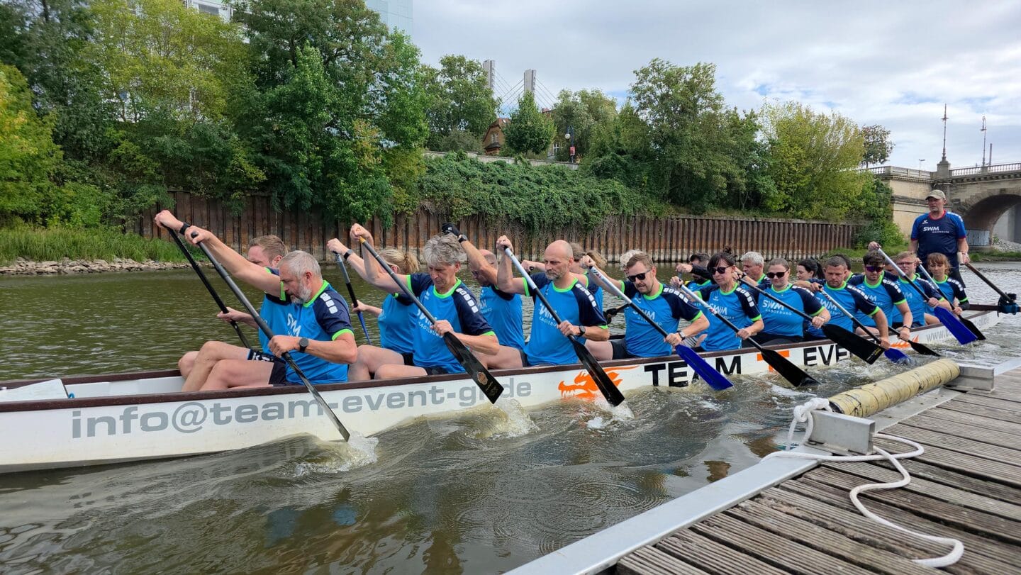 Ein großes weißes Boot mit mehreren Menschen in blauen Trikots die auf einem Fluss paddeln.