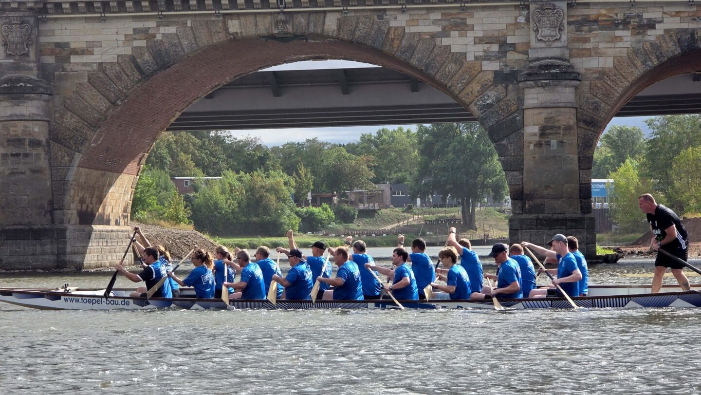 Ein großes weißes Boot mit mehreren Menschen in blauen Trikots die auf einem Fluss paddeln.