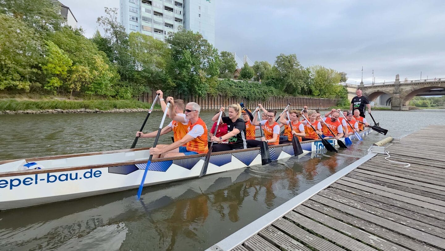 Ein großes weiße Boot mit mehreren Menschen in orangen Trikots die auf einem Fluss paddeln.