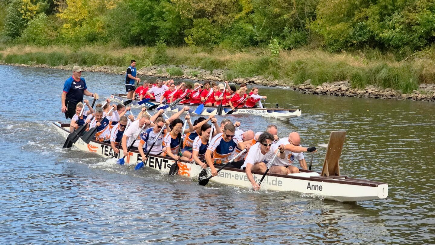 Zwei große weiße Boot mit mehreren Menschen in weißen und roten Trikots die auf einem Fluss angestrengt paddeln.