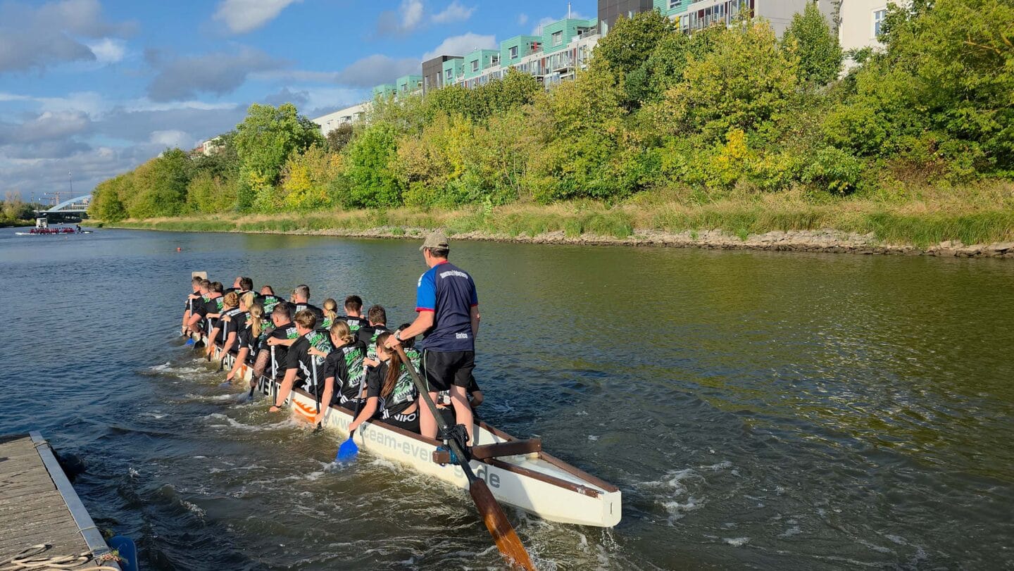 Ein großes weißes Boot mit mehreren Menschen in schwarzen Trikots die auf einem Fluss paddeln.