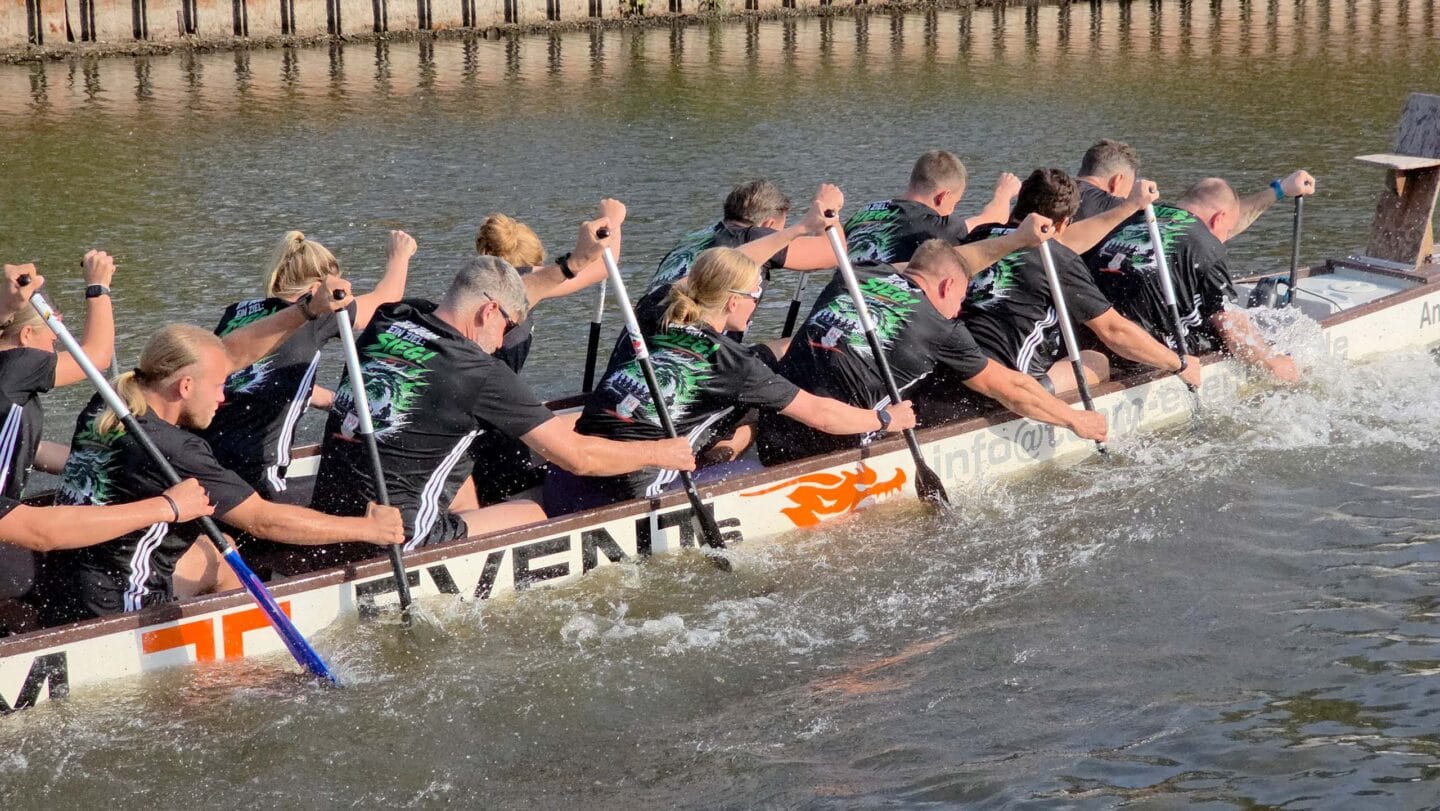 Ein großes weißes Boot mit mehreren Menschen in schwarzen Trikots die auf einem Fluss angestrengt paddeln.