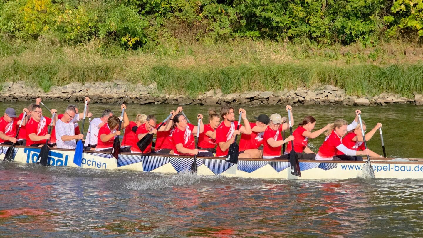 Ein großes weißes Boot mit mehreren Menschen in roten Trikots die auf einem Fluss paddeln.