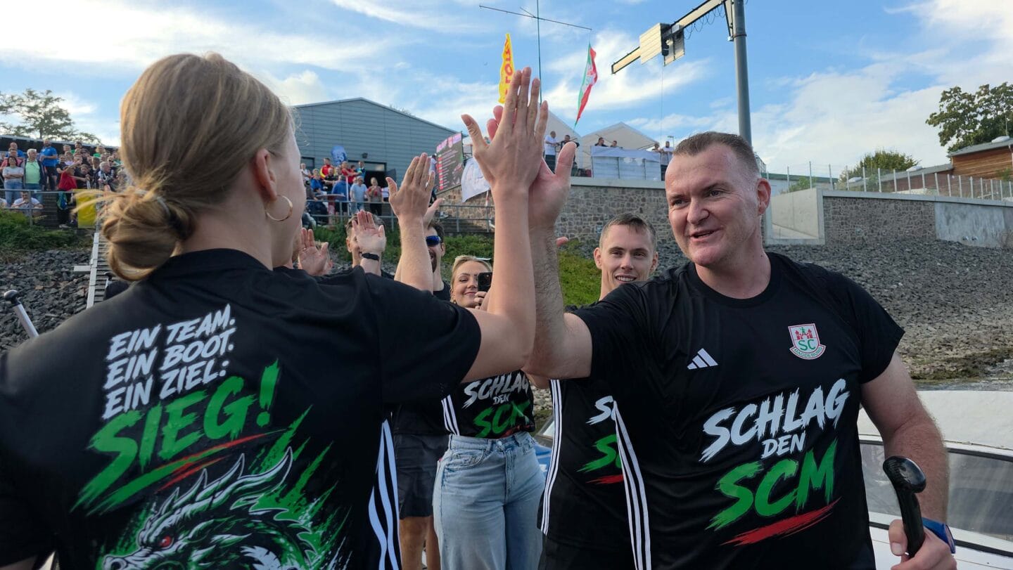 Mann im schwarzen T-Shirt klatscht mit einer Frau in einem schwarzen T-Shirt ab. Dahinter weitere Personen auf einem Bootssteg.