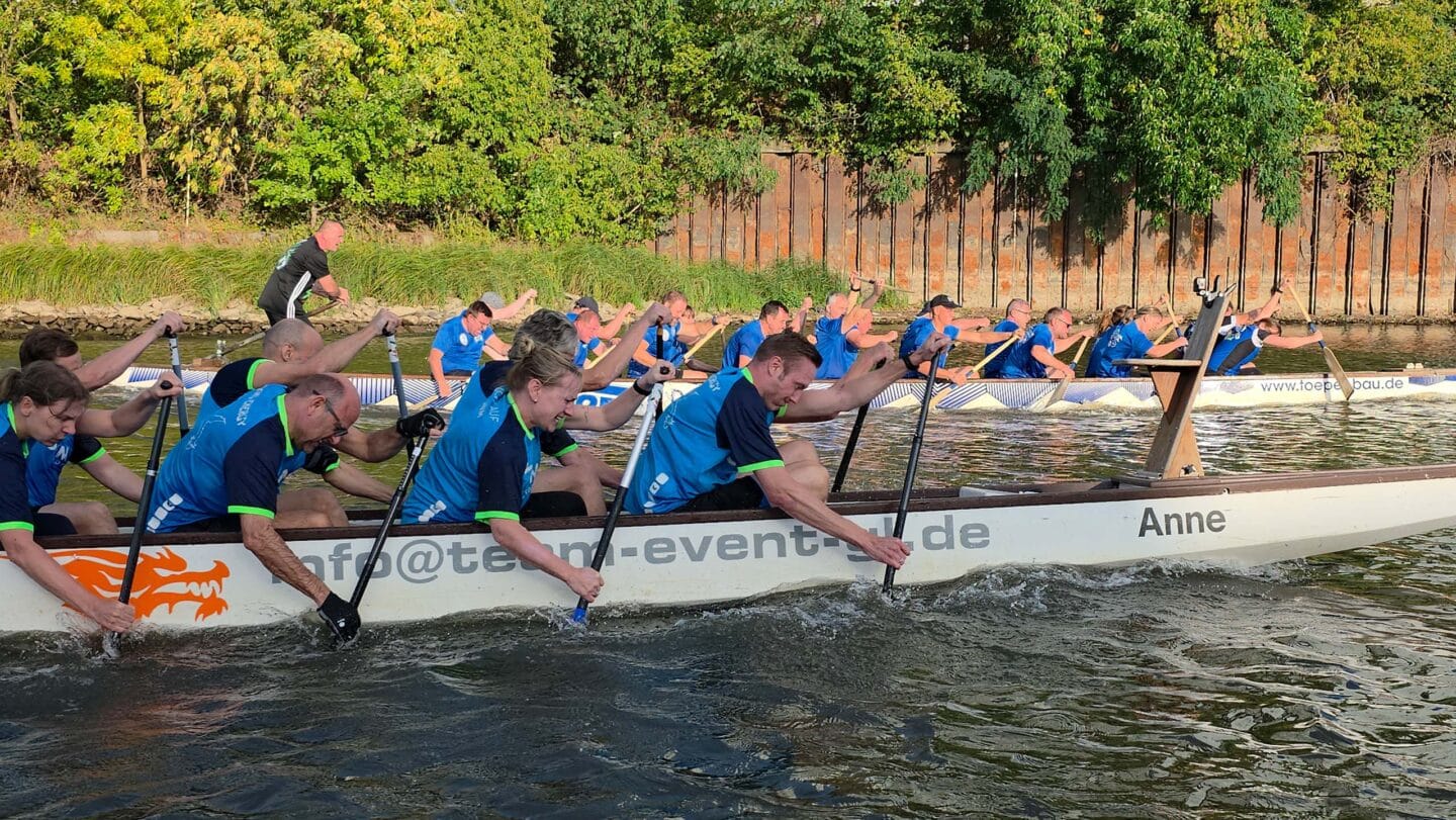 Zwei große weiße Boot mit mehreren Menschen in blauen Trikots paddeln angestrengt auf einem Fluss.