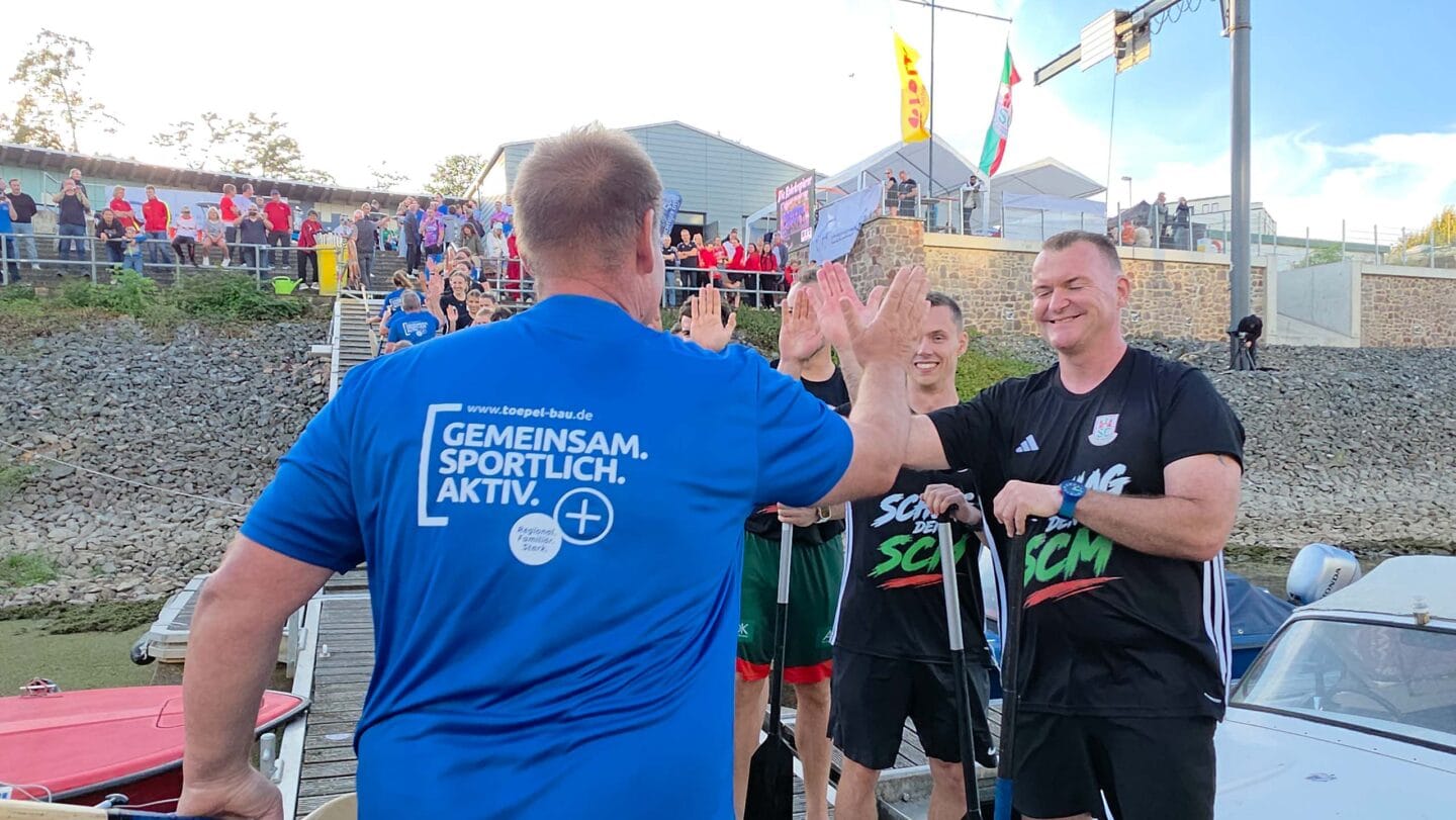Mann im blauen T-Shirt klatscht mit einem Mann in einem schwarzen T-Shirt ab. Dahinter weitere Personen auf einem Bootssteg.
