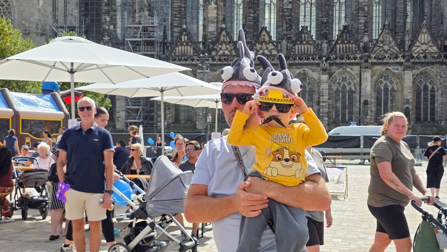 Ein Mann mit einem kleinen Kind auf dem Arm. Das kind trägt eine gel-oranges Shirt auf dem ein Hund ist. Beide tragen eine Sonnenbrille und Haisfisch-Mützen.