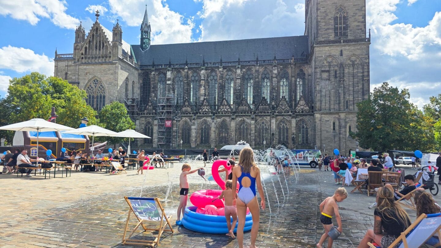 Wasserfontänen auf einem gepflasterten Platz mit bunten kleinen Planschbecken in den Kindern mit Badesachen spielen. Im Hintergrund der Magdeburger Dom.