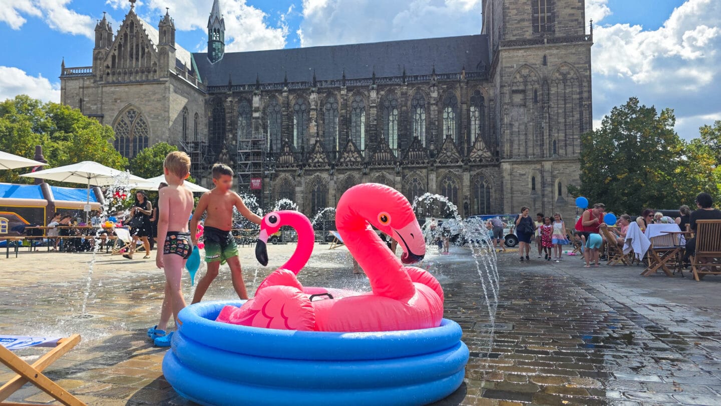 Wasserfontänen auf einem gepflasterten Platz mit bunten kleinen Planschbecken in den Kindern mit Badesachen spielen. In den Planschbecken liegen rosa Plastik-Flamingos.