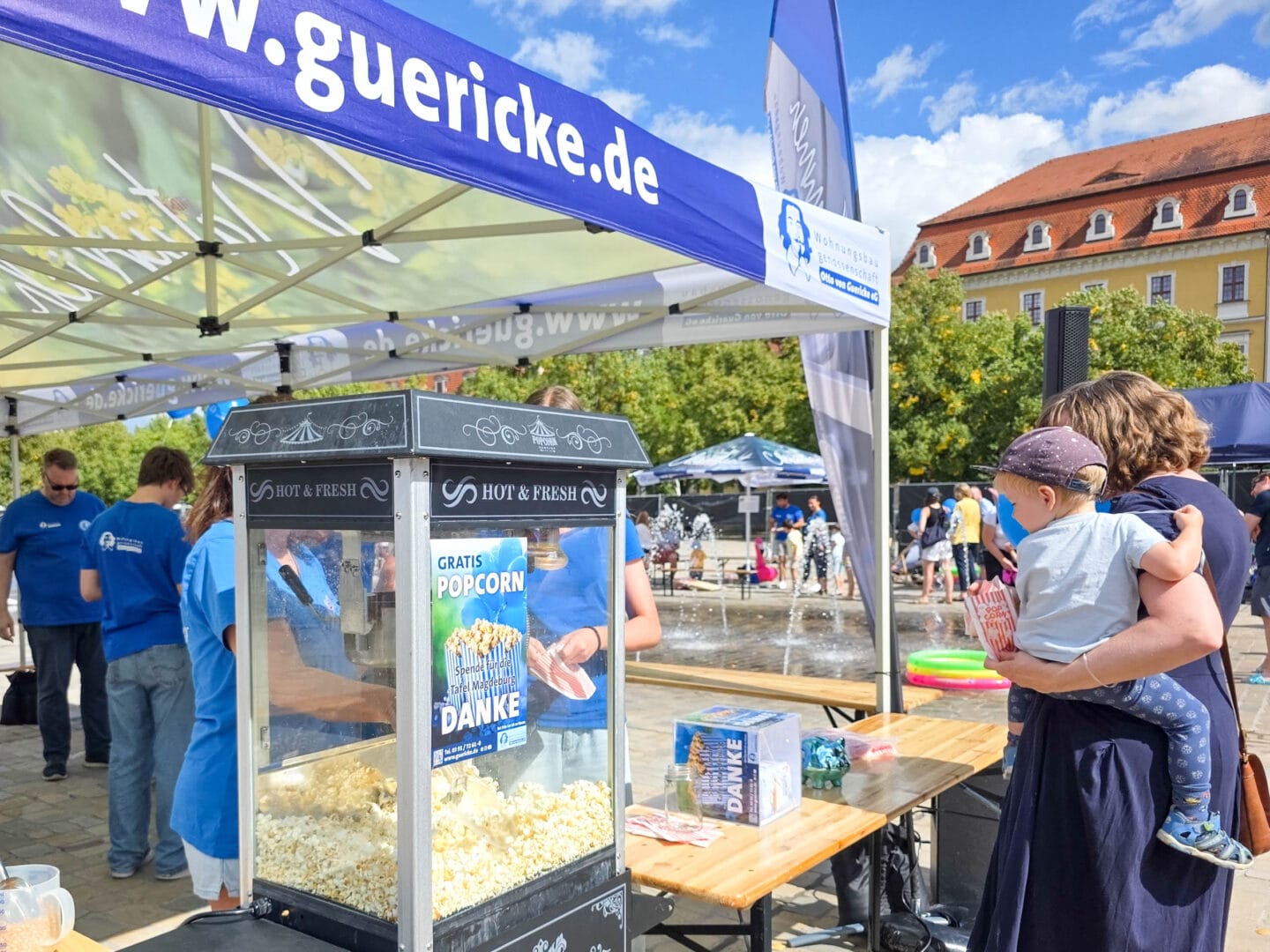Eine Popcorn-Maschine aus Glas mit viel Popcorn und davor stht eine Frau mit einem KInd auf dem Arm. Im Hintergrund mehere Menschen in blauen T-Shirts.