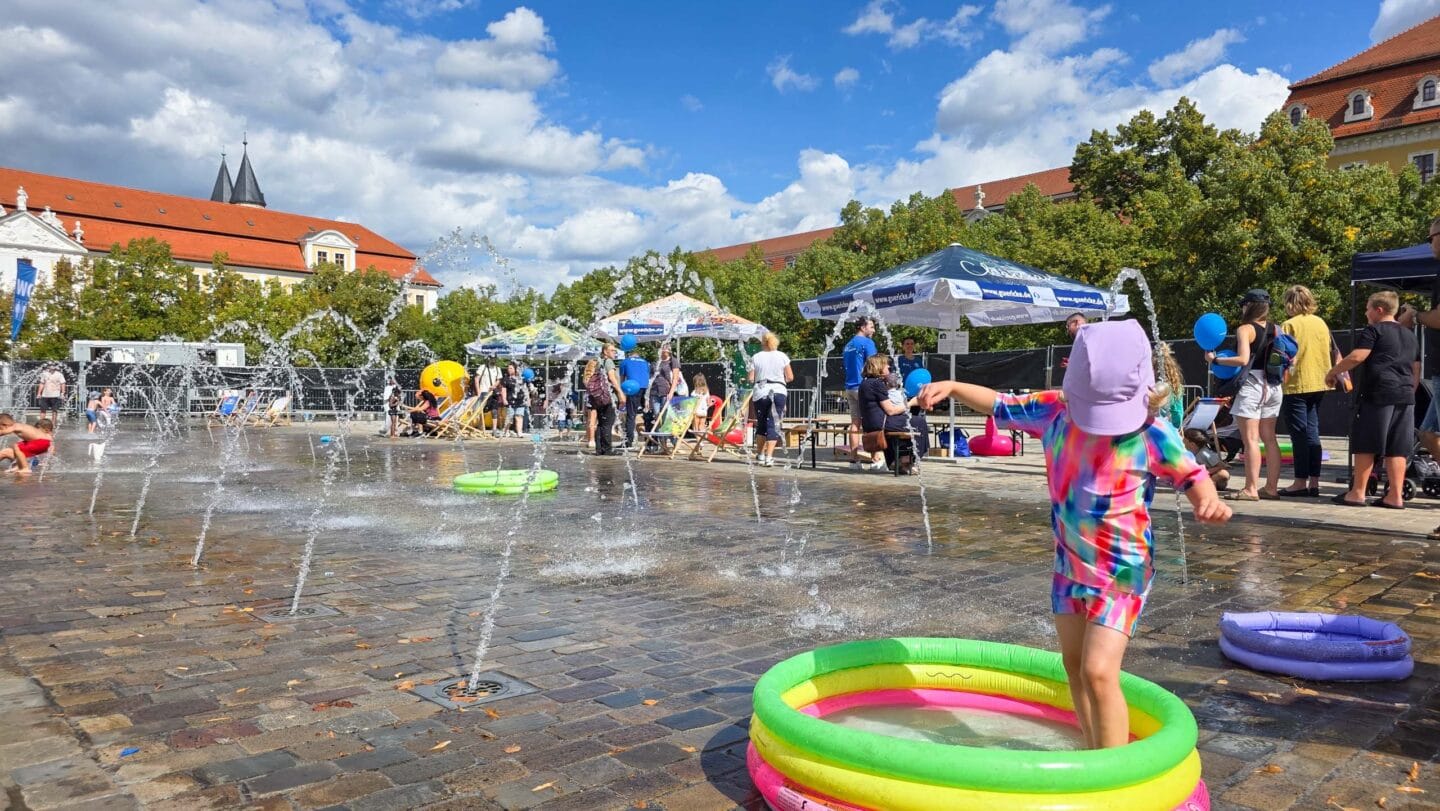 Wasserfontänen auf einem gepflasterten Platz mit bunten kleinen Planschbecken in dem ein Kinder mit Badesachen spielet.