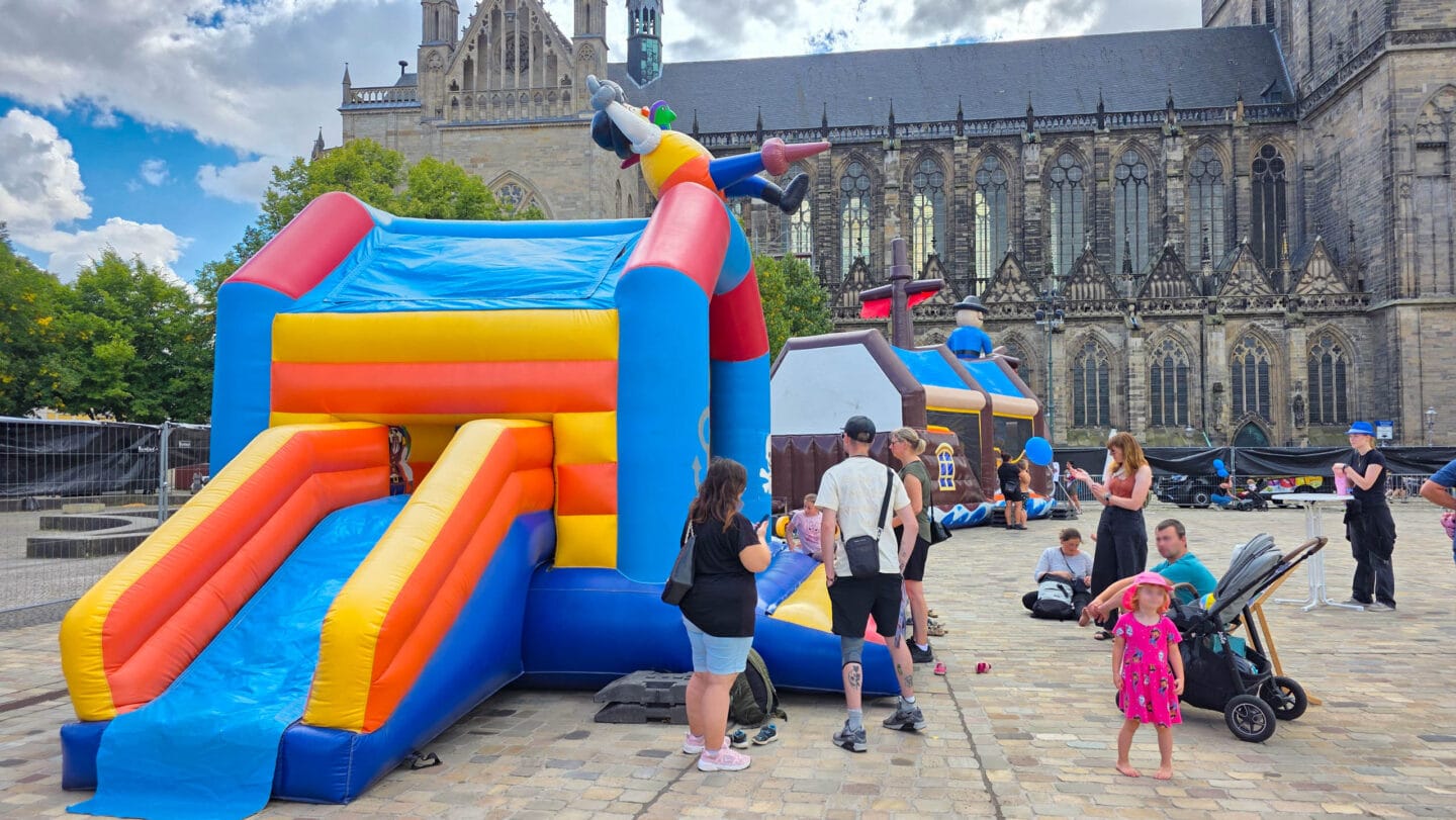Eine große Hüpfeburg in der Form eines bunten Hauses und dahinter in Form eines Priratenschiffs auf dem Domplatz. Davor stehen mehere Frauen und Männer