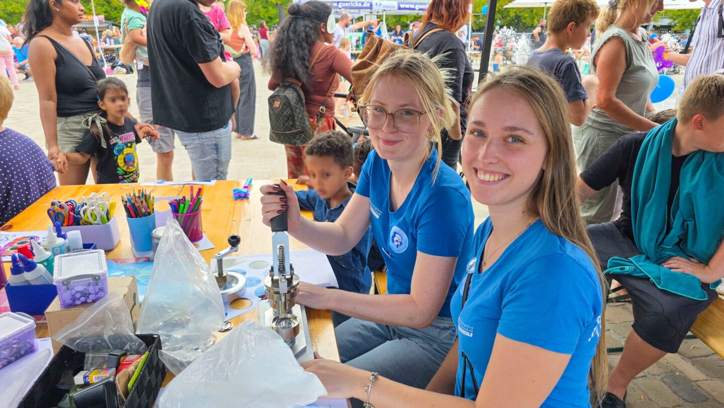 Zwei junge blonde Frauen in blauen T-Shirts sitzen an einem Tisch mit Bastelsachen und lächeln in die Kamera.