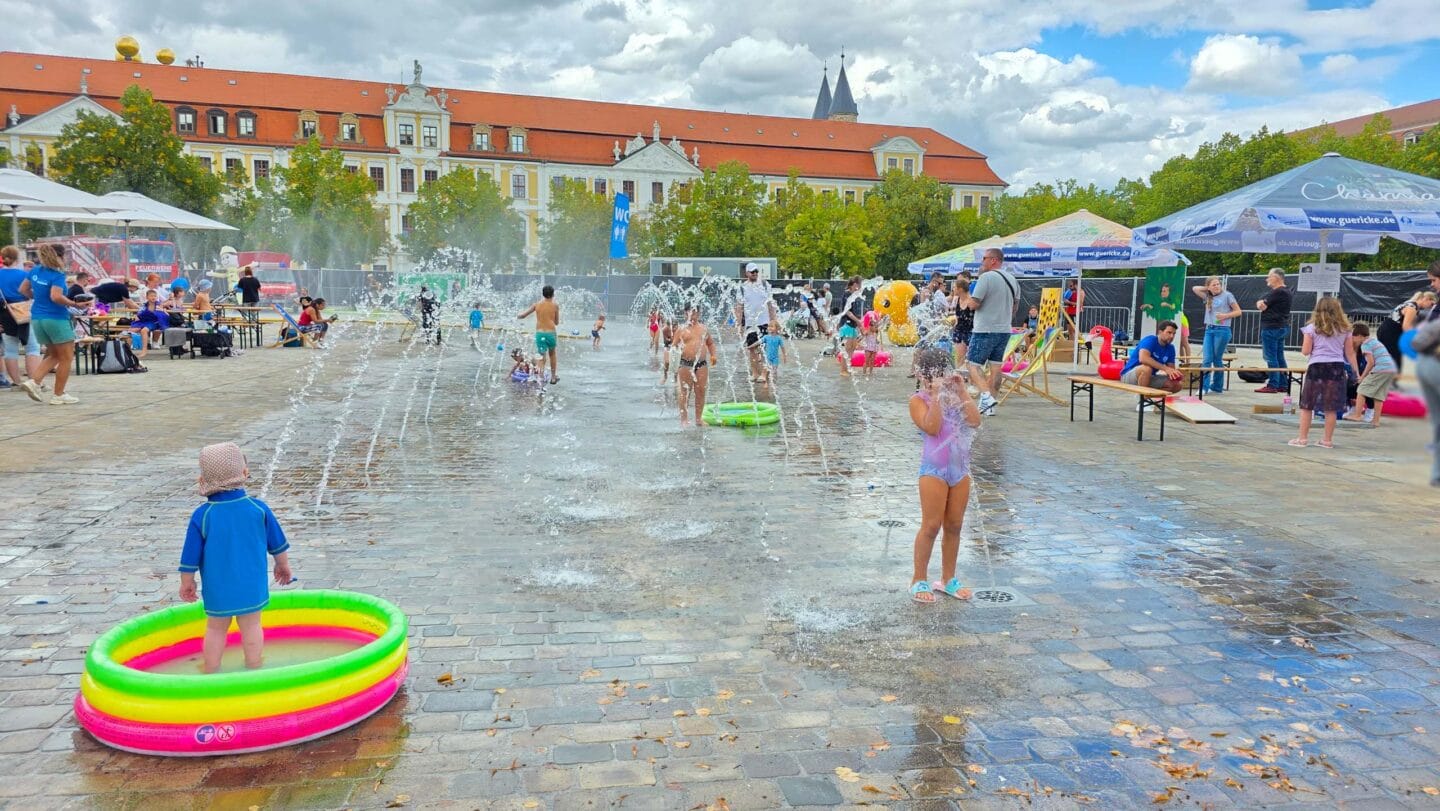 Wasserfontänen auf einem gepflasterten Platz mit bunten kleinen Planschbecken in den Kindern mit Badesachen spielen.