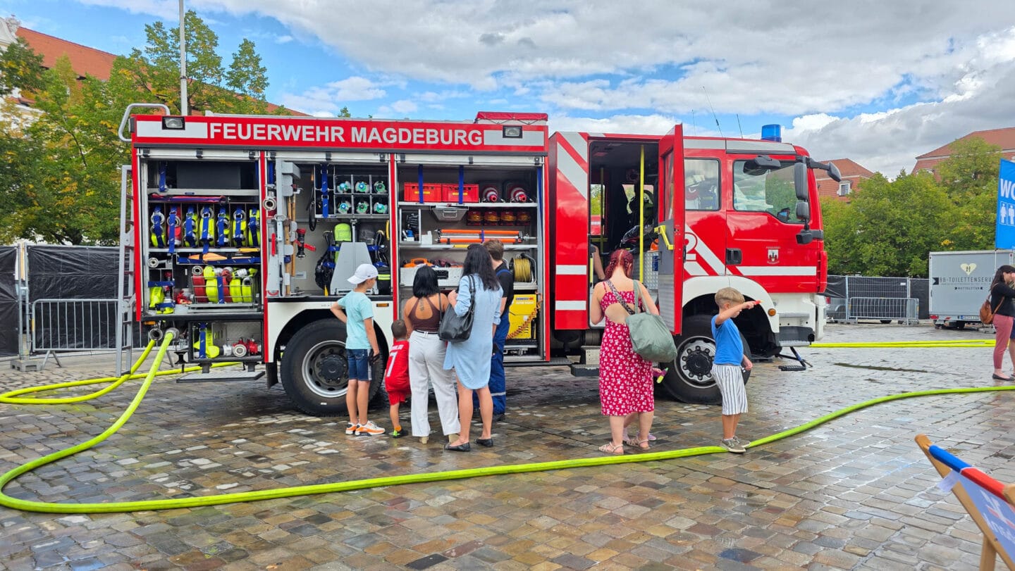 Ein großes rotes Feuerwehraout mit offenene Türen und davor mehere Menschen in sommerlicher Kleidung