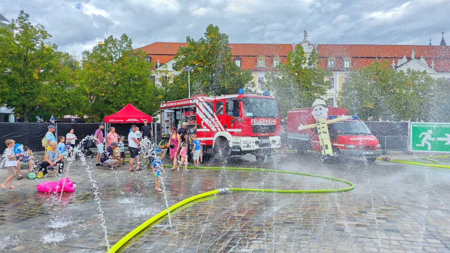 Wasserfontänen aus neongelben Wasserschläuchen. Dahinter ein großes rotes Feuerwehrauto mit mehreren Menschen.