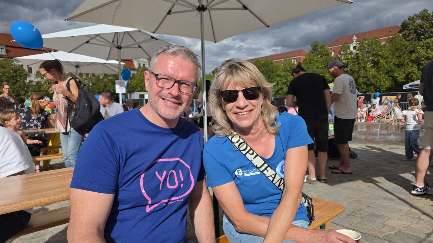 Vorstände Oliver Hornemann und Karin Grasse lächeln in blauen T-Shirts in die Kamera. im Hintergrund Sonnenschirme und mehere Menschen