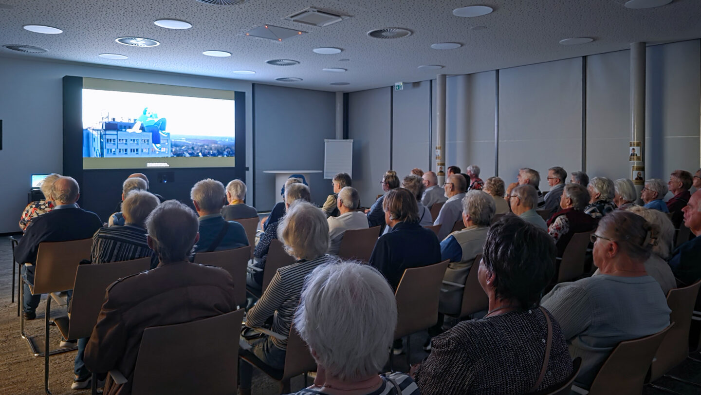 Eine Gruppe von älteren Menschen sitzen und schauen auf eine Leinwand.