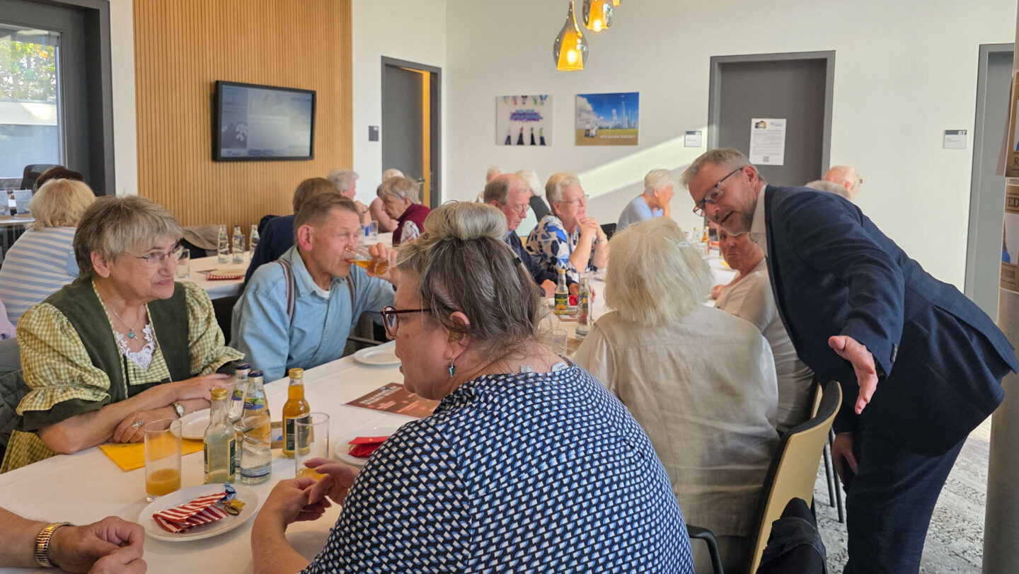 Eine Gruppe Menschen sitzen am Tisch und unterhalten sich. Ein Mann im Anzug steht dahinter und unterhält sich.