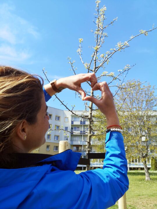 Frau formt aus ihren Händen ein Herz und deutet zum frisch gepflanzten Baum. ©Anja Jänichen