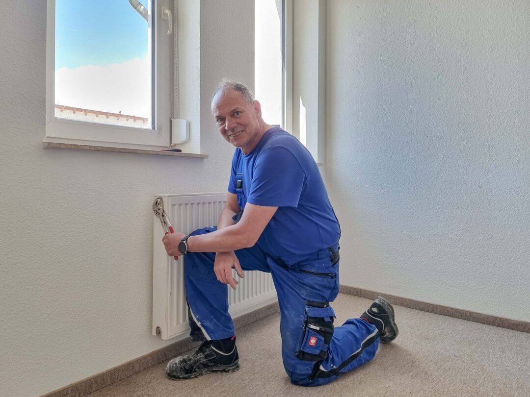 Ein Mann mit blauer Arbeitskleidung kniet vor einer Heizung, repariert si und lächelt in die Kamera.