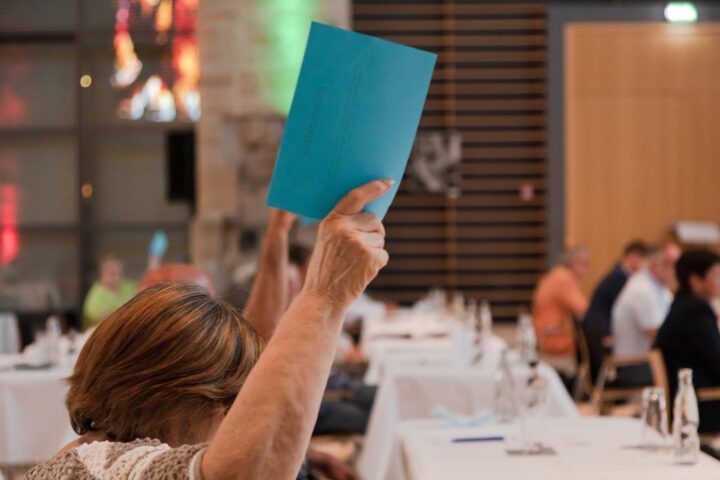 Eine Frau hält einen blauen Zettel hoch und sitzt an langem Tisch mit anderen Menschen. Ihr Gesicht ist nicht zu erkennen.